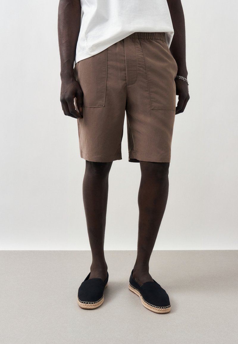 Άτομο που φοράει καφέ σορτς μέχρι το γόνατο, λευκό κοντομάνικο, μαύρα εσπαντρίγιες και ασημένιο βραχιόλι, στέκεται μπροστά σε μια απλή ανοιχτή φόντο.