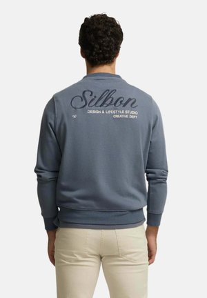 Hombre con pelo rizado oscuro que lleva una sudadera azul con el texto "Silbon Design & Lifestyle Studio Creative Dept" en la espalda y pantalones beige.