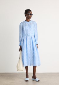 Light blue polka dot dress with long sleeves, button-down front, belted waist, and a flowy A-line shape. Worn with light blue mules and a cream bag.