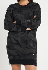 Mustat kamouflage-puseromekot, joissa on tekstuuri, pyöreä pääntie, pitkät hihat ja musta resori. Sivuvetoketjut ja logoyksityiskohta.
