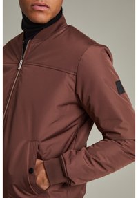 Brun bomberjacka i slätt tyg med dragkedja framtill, sidfickor, ribbade muddar och en svart detalj på ärmen.