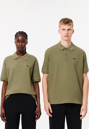 Camisetas polo verde oliva con manga corta, tapeta de doble botón y logotipo de cocodrilo bordado. Llevadas con pantalones negros.