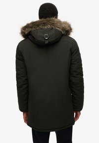 Opgestikte zwarte buitenjas met een met fauxbont gevoerde capuchon en verstelbare band, met een subtiele logo op de achterkant. Lange mouwen en een rechte snit.