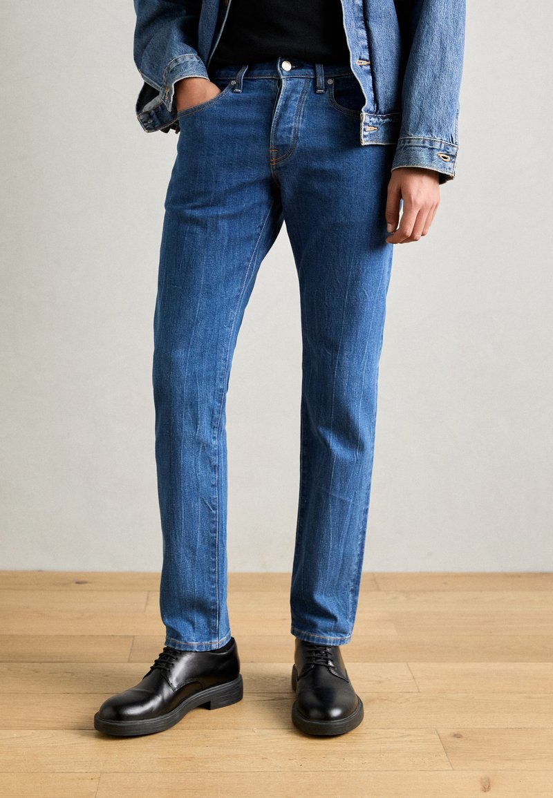 Blå denimjeans med slim fit, der har subtile falmning detaljer. Sat sammen med sorte læder snørebåndssko på et trægulv.