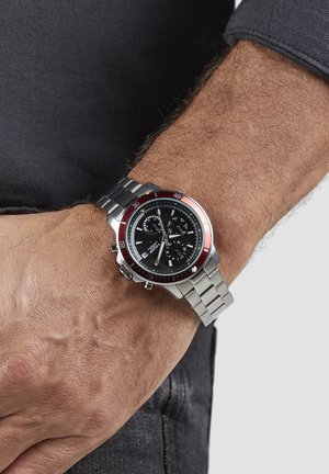 Montre chronographe avec un bracelet en acier inoxydable, un cadran noir, une lunette rouge, des accents argentés et trois sous-cadrans. Portée sur un poignet avec des poils visibles.