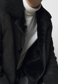 Manteau en shearling noir avec un col en fausse fourrure, orné de boutons, superposé à un pull à col roulé blanc. Texture en cuir lisse, design angulaire.