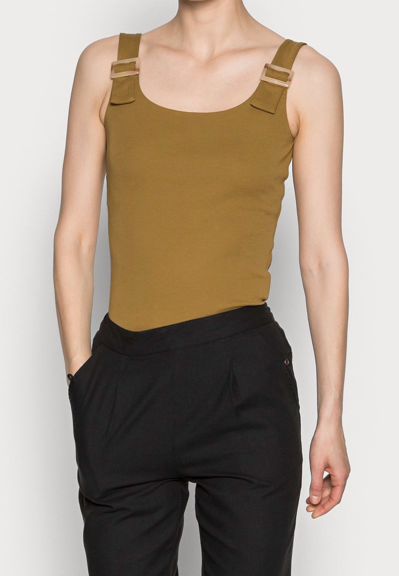 Personne portant un haut sans manches vert olive avec des bretelles à boucle et un pantalon noir, les mains dans les poches.