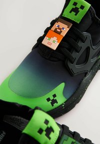 Sneaker neri e verdi con una parte superiore a gradiente, caratterizzati da accenti di personaggi pixelati e una suola testurizzata con un elemento di design giocoso.