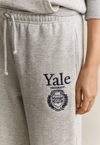 Ανοιχτόγριμο φόρμας με το λογότυπο του Yale University κεντημένο σε σκούρο μπλε στο αριστερό μηρό, που δείχνει μέρος του χεριού και το ταιριαστό τοπ από το ίδιο ύφασμα.