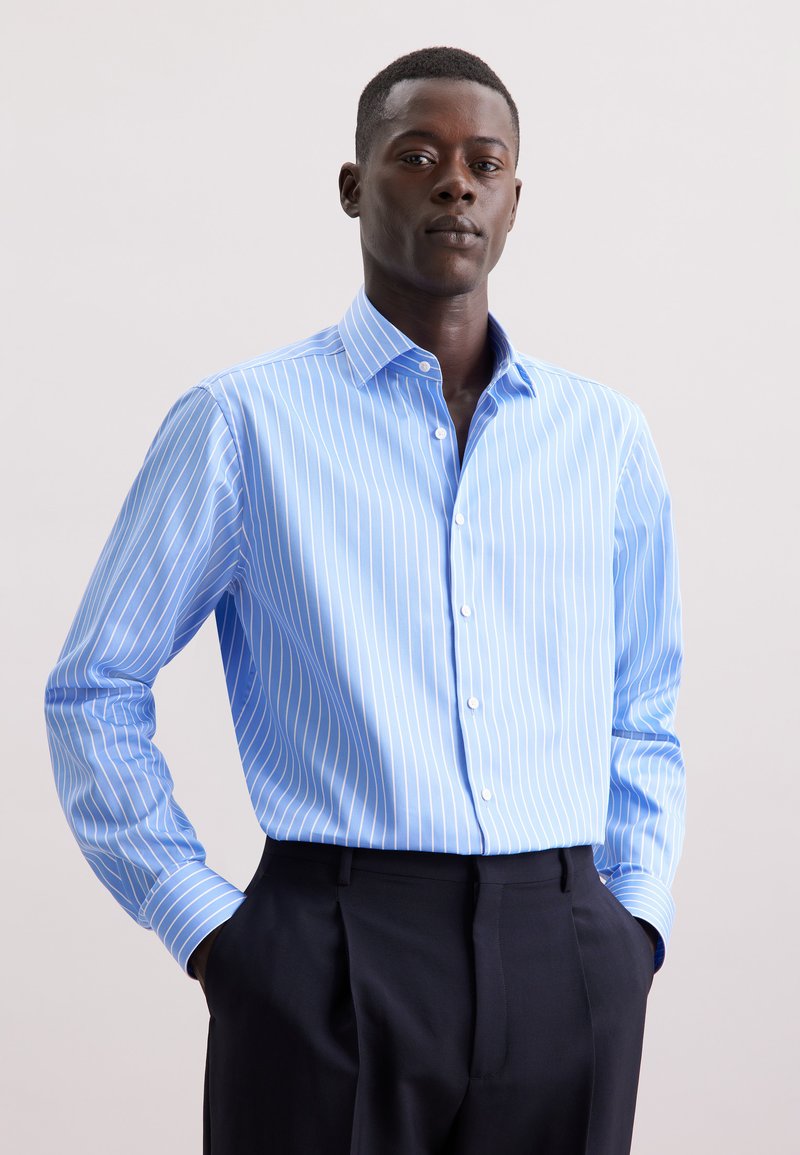 Camicia button-up azzurro chiaro con sottili strisce verticali bianche, maniche lunghe e colletto classico. Indossata con pantaloni scuri.