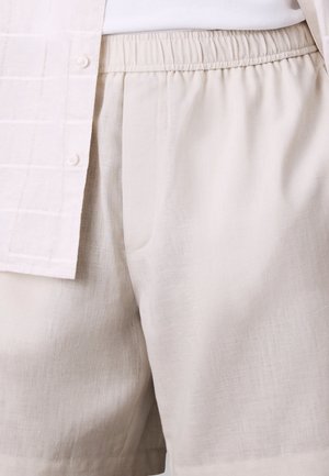 Pantaloncini di lino beige chiaro con elastico in vita abbinati a una camicia bianca infilata con sottili motivi a quadretti.