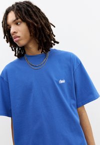 Blå bomulds T-shirt med korte ærmer, der har et lille hvidt logo på venstre bryst og en afslappet pasform. Glat tekstur, enkelt design.
