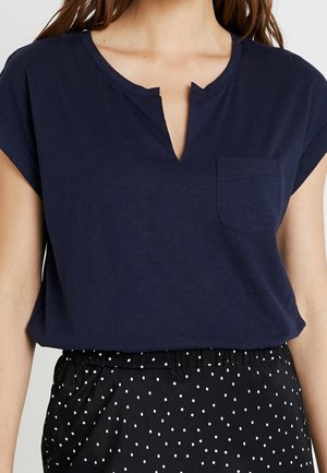 Marineblaues Kurzarmoberteil aus weichem Stoff mit V-Ausschnitt und Taschenverzierung, kombiniert mit schwarzen Hose mit Tupfenmuster und elastischem Bund.