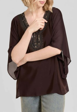 Donna con camicetta ampia marrone scuro con scollo a V decorato e maniche larghe, braccia incrociate, indossa jeans azzurri chiaro su sfondo neutro.