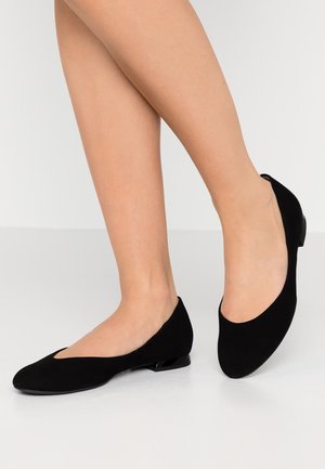 Jambes portant des ballerines noires en suède à talons bas et bouts arrondis, posées sur un sol gris clair avec un fond blanc.