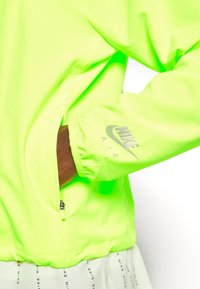 Persona che indossa una giacca Nike Air verde neon con una mano nella tasca con zip, gonna bianca Nike con testo nero visibile sotto la giacca.