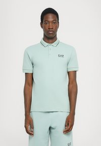 Polo de cor verde claro com mangas curtas, colarinho e costura de contraste preta, apresentando um pequeno logotipo preto no peito.