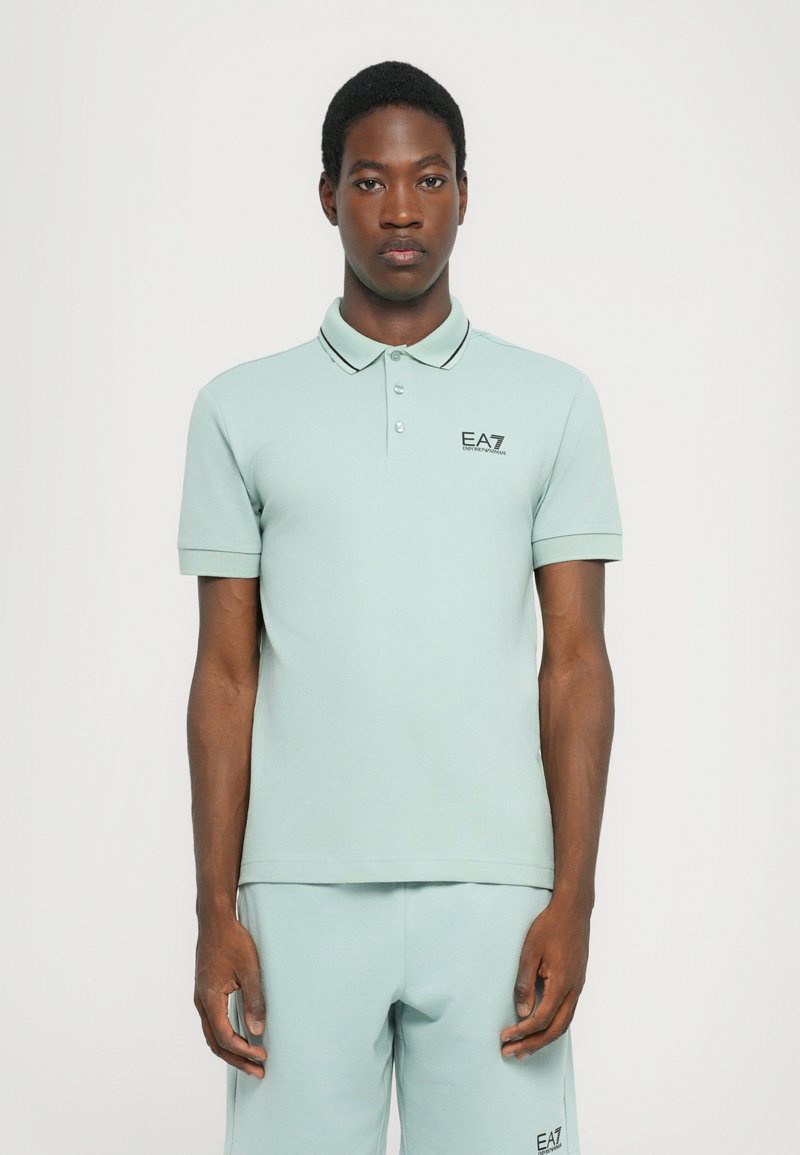 Polo de cor verde claro com mangas curtas, colarinho e costura de contraste preta, apresentando um pequeno logotipo preto no peito.