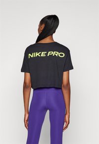 Sort cropped t-shirt har "NIKE PRO" i fed, reflekterende neongul tekst på ryggen, parret med glatte lilla leggings.