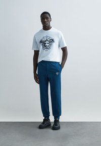 Vit bomullst-shirt med blå Medusa-grafik och texten "Versace". Blå byxor med broderad logotyp, matchade med svarta sneakers.