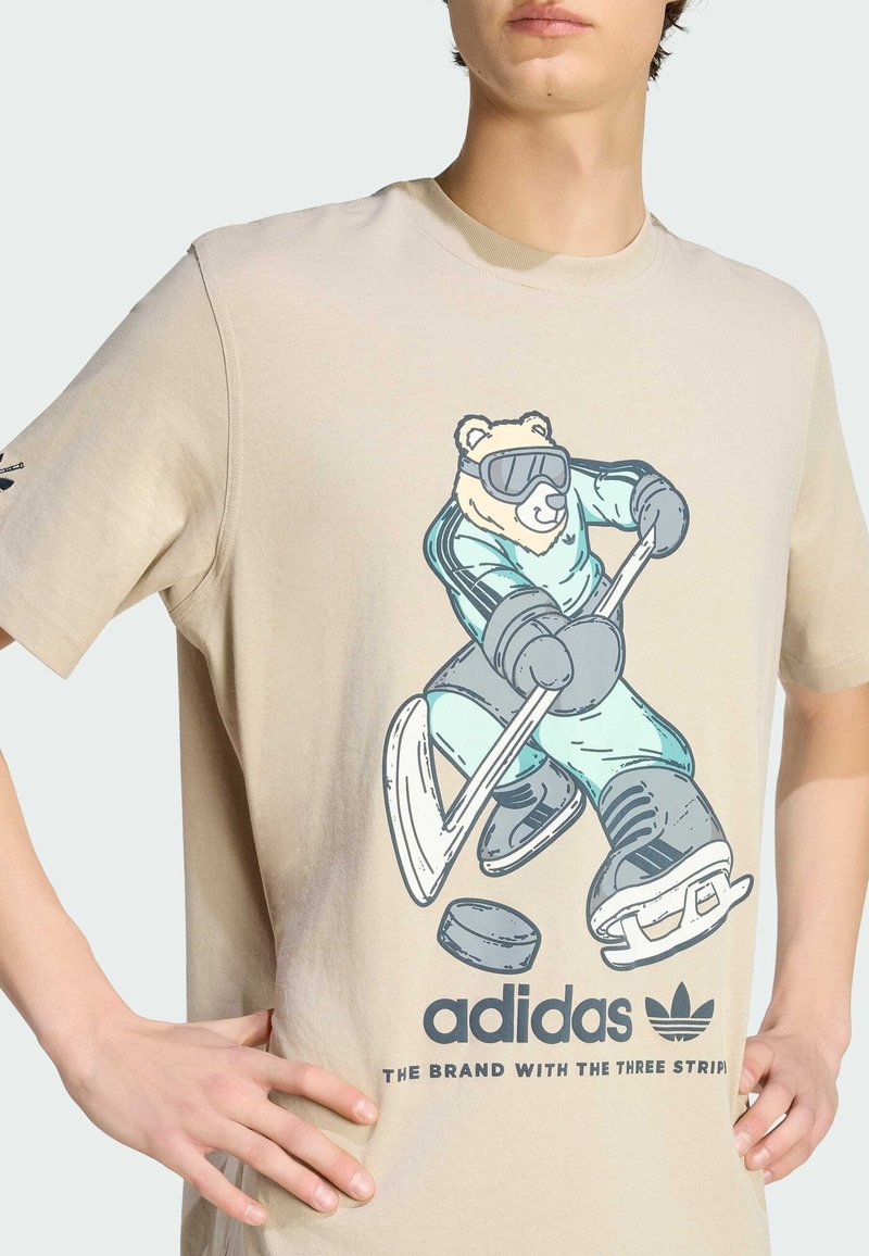 Maglietta beige in cotone con un orso dei cartoni animati in tenuta da sci che tiene una stecca da hockey, con il logo "adidas" stampato sotto.