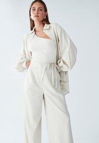 Svetlo bež oversize majica z zavihanimi rokavi, v kombinaciji s hlačami širokega kroja z visokim pasom in vezalko. Teksturovana, mehka tkanina.