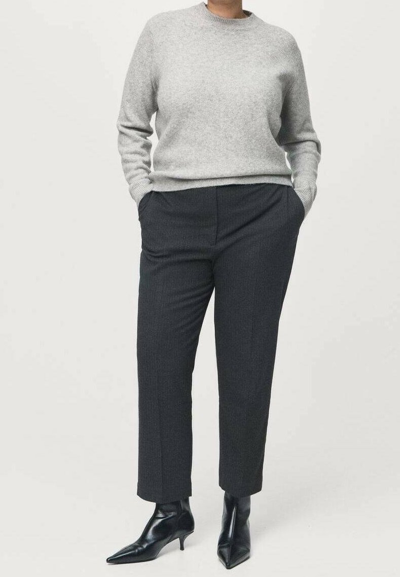 Personne portant un pull gris clair, un pantalon tailleur gris foncé taille cheville, et des bottines noires à talons pointus, debout les mains dans les poches.