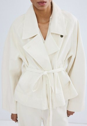 Person wearing an off-white belted jacket with wide lapels and matching off-white pants, standing against a plain background.