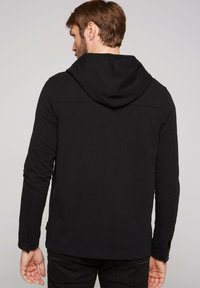 Giacca nera con cappuccio e maniche lunghe, realizzata in un tessuto liscio, con dettagli a bottoni ai polsini e un design semplice ed essenziale.