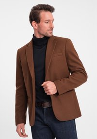 Brun ullblandad kavaj med slag och en enkel bröstficka, lager på lager över en svart polotröja, kombinerad med blå denimjeans.