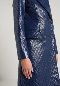 Giacca trapuntata blu navy realizzata in materiale lucido, caratterizzata da un design doppiopetto con due bottoni scuri e una vestibilità su misura.