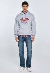 Ein junger Mann steht da und trägt einen grauen Hoodie mit "Vintage"-Druck, ein kariertes Hemd darunter, blaue Jeans und weiße Turnschuhe vor einem einfarbigen Hintergrund.