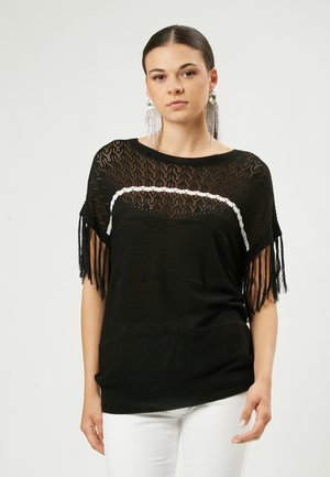 Top de punto negro con diseño de encaje abierto, franja de acento blanca, mangas cortas y bordes con flecos. Combinado con pantalones blancos, creando un efecto de capas.