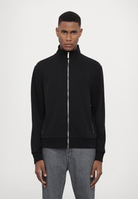 Giacca nera con zip e colletto alto, realizzata in materiale morbido. Presenta una superficie liscia e tasche laterali, abbinata a jeans grigi.