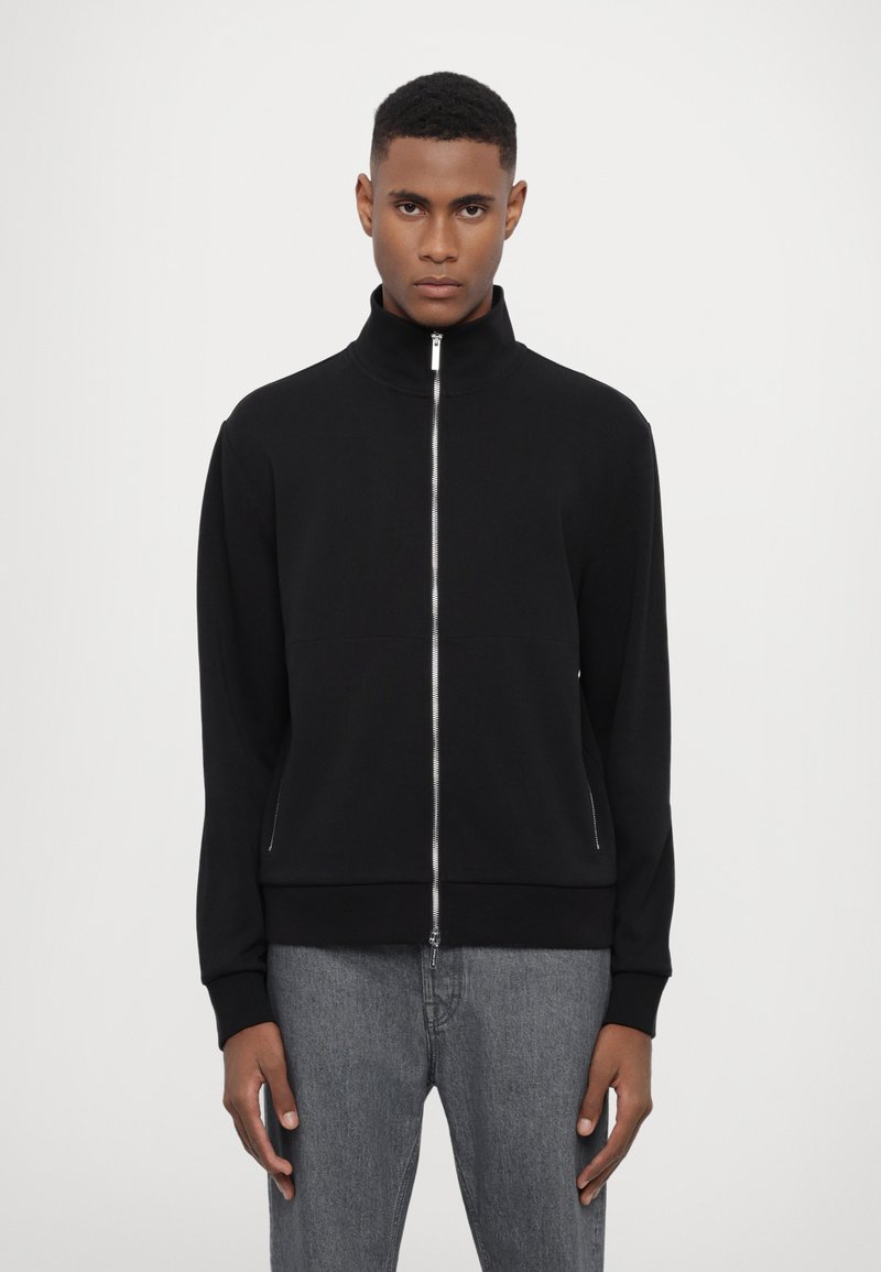 Giacca nera con zip e colletto alto, realizzata in materiale morbido. Presenta una superficie liscia e tasche laterali, abbinata a jeans grigi.