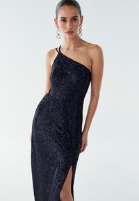 Námorníckej modrej večernej šaty so asymetrickým výstrihom a vysokým rozparkom. Obsahuje trblietavú textúru a úzky strih, ktorý zvýrazňuje siluetu.