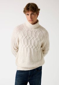 Maglione chunky lavorato a maglia di colore bianco sporco con collo alto a funnel. Presenta un motivo in rilievo a treccia e orli a coste sia in fondo che sui polsini. Abbinato a jeans scuri.
