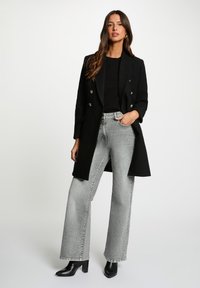 Sort dobbeltradet frakke med sølvknapper, båret over en sort top, grå bukser med bredt ben og sorte højhælede støvler. Glat tekstur, skræddersyet pasform.