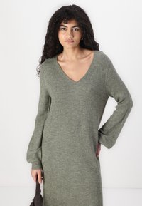 Robe tricot verte avec un décolleté en V et des manches longues. Elle présente une texture douce et une coupe ample, atteignant la longueur mi-mollet.