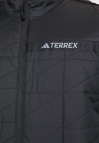 Czarna izolowana kurtka z pikowanym wzorem, z przodem na zamek błyskawiczny oraz szarym logo "TERREX" na poziomym pasie. Gładka tekstura i lekkość.