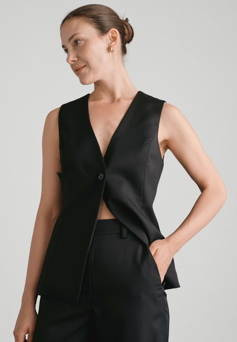 Czarny bezrękawnik z głębokim dekoltem w kształcie V, pojedynczym zapięciem na guzik, usztywnioną konstrukcją i luźnym krojem. Gładka tkanina o subtelnym połysku.
