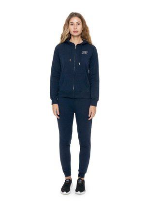 Une femme porte un sweat à capuche bleu marine zippé et un pantalon de jogging assorti, avec des baskets noires, sur fond blanc.