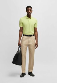 Ljusgrön pikétröja, beige byxor, svart bälte och skor. Håller en svart väska. Kort ärm och klassisk kragsdesign.