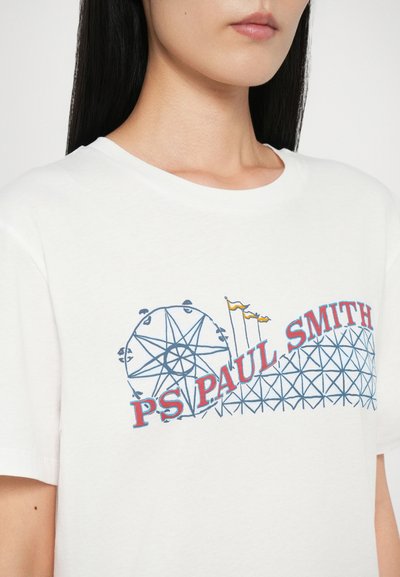 Balts kokvilnas T-krekls ar grafisku dizainu, kur attēlots rīģu ritenis un kalniņš, ar "PS PAUL SMITH" tekstu sarkanā un zilā krāsā.