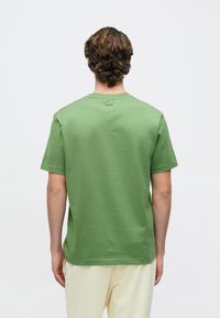 Πράσινο βαμβακερό T-shirt με κοντά μανίκια, στρογγυλή λαιμόκοψη και υφή στην επιφάνεια. Μικρή λεπτομέρεια λογότυπου στο πάνω μέρος της πλάτης. Ουδέτερο φόντο.