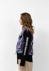 Paarse fleece vest met een luipaardprint; zijzak met rits; gedragen over een zwart longsleeve shirt met witte tekst op de mouw.