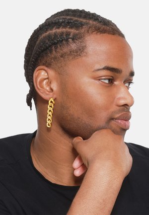 Boucle d'oreille style chaîne en or avec des maillons interconnectés, portée à l'oreille gauche. Gros plan sur le visage, peau lisse, portant une chemise noire.