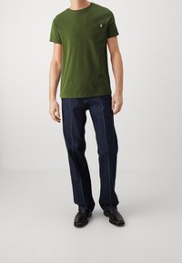 Camiseta de algodón verde oliva con bolsillo en el pecho, combinada con vaqueros oscuros con costuras azules y botas de cuero negras.