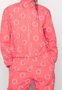 Korallrosa träningsdräkt med en zip-up jacka och byxor med elastisk midja, prydd med upprepade vita cirklar med "adidas"-logotyp.