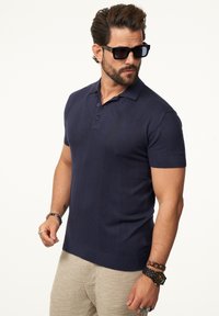Marineblaues Polohemd mit kurzen Ärmeln, texturiertem Ripp-Paneel, Knopfleiste mit drei Knöpfen und taillierter Passform, kombiniert mit hellen, texturierten Hosen.